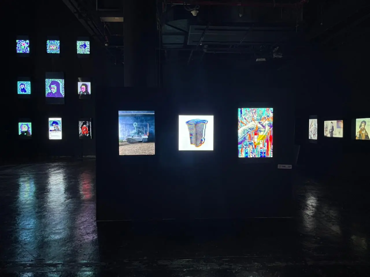 A dark space with a freestanding wall of screens and a grid of works behind